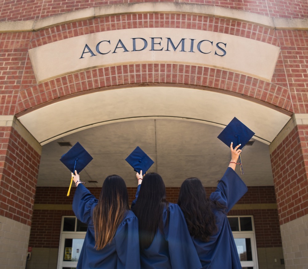Three graduates in blue gowns and caps raise their hands under a brick archway with the inscription 