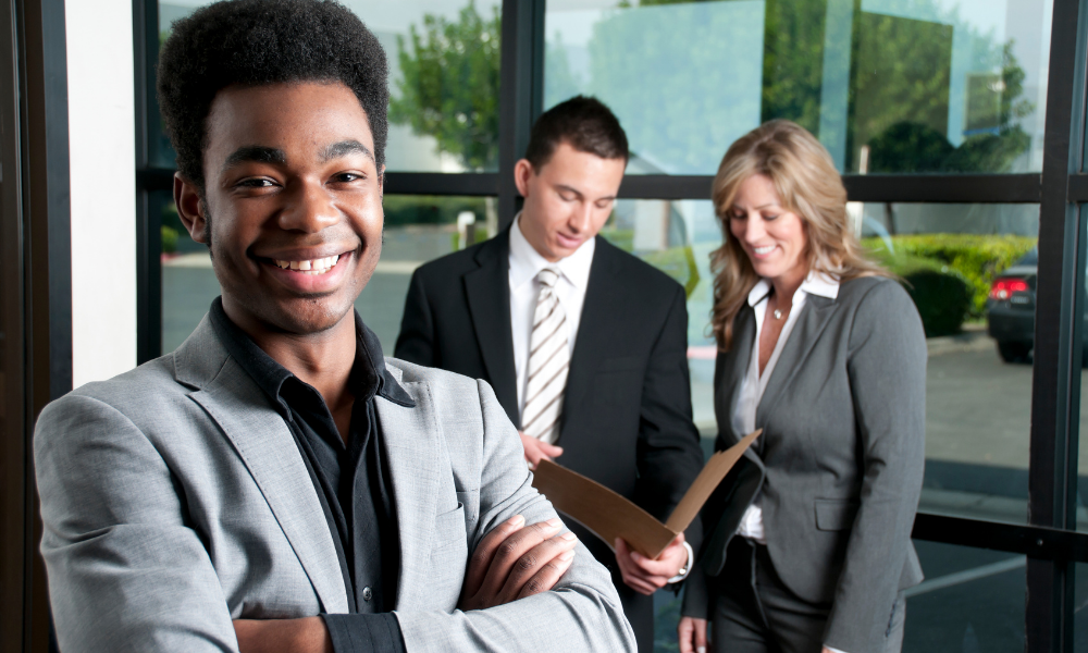 Three graduate hires stand in front of large windows, with one in the foreground smiling at the camera while the other two discuss a document in the background.