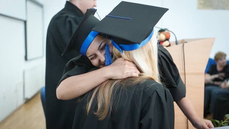 Two graduates in black caps and gowns, displaying exemplary people management skills, hug each other during a graduation ceremony. One has a blue tassel hanging off the cap, marking an unforgettable milestone in their personal and professional journey.