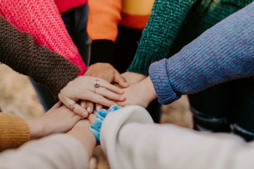 A group of people stand in a circle, placing one hand each in the center on top of one another, showing unity and teamwork amidst the great resignation. They are wearing colorful sweaters.