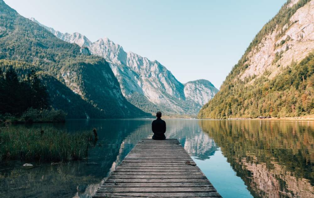 A person sits at the end of a wooden dock facing a calm lake, surrounded by mountains and greenery under a clear sky, immersed in transcendental meditation.