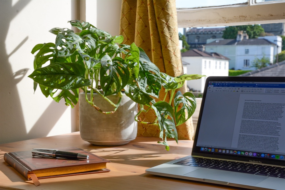 A laptop displaying a document is on a wooden desk next to a notebook, pens, and a potted plant. Sunlight shines through a window with yellow curtains, creating the perfect homeworking environment.