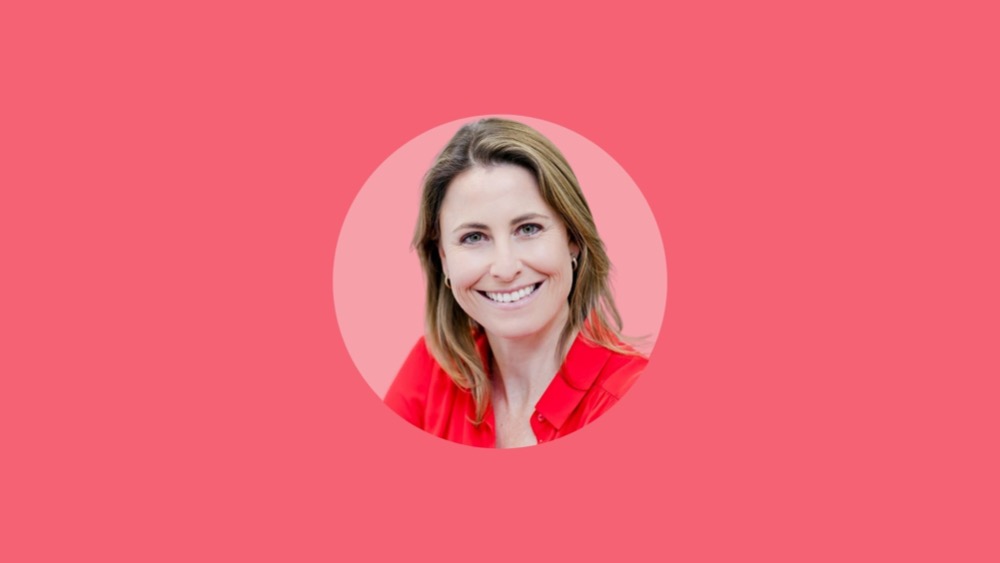 A woman with shoulder-length hair smiles, wearing a red top. She stands confidently against a solid pink background, representing the modern shift to hybrid working.