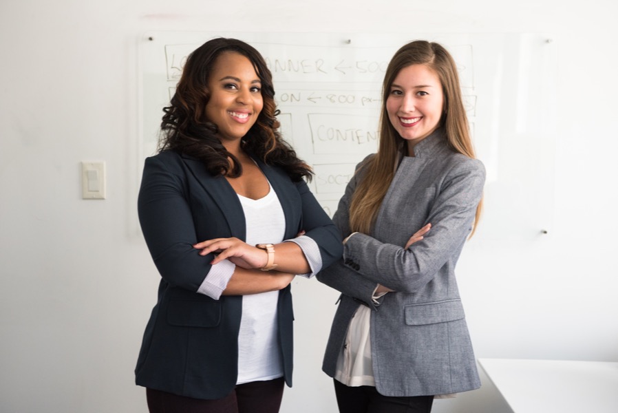 Two women standing with arms crossed in front of a whiteboard in an office setting, both wearing blazers and smiling confidently at the camera, showcasing a team dynamic that thrives without preferential treatment.