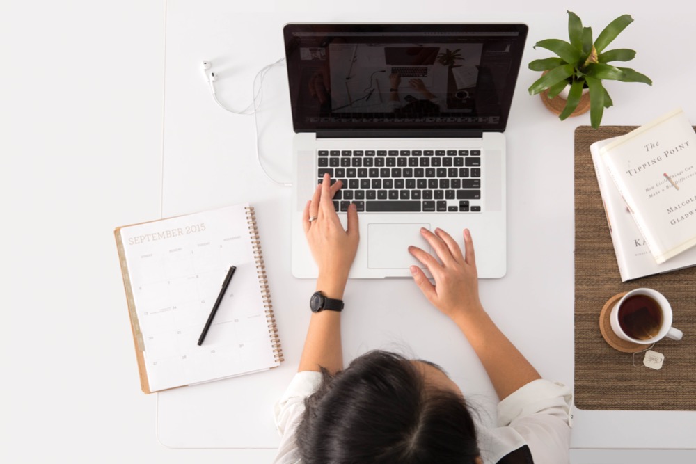 A person engaged in remote working sits at a desk with a laptop, calendar, pen, plant, book, and a cup of coffee.
