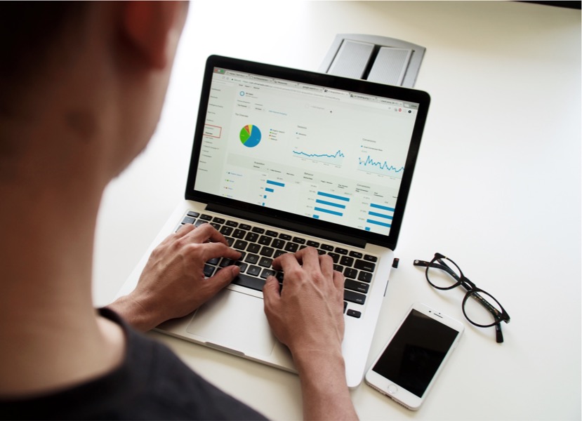 An employee is working on a laptop displaying graphs and charts, with a smartphone and eyeglasses placed beside the laptop on a white desk, epitomizing the drive for employee engagement through effective data analysis.