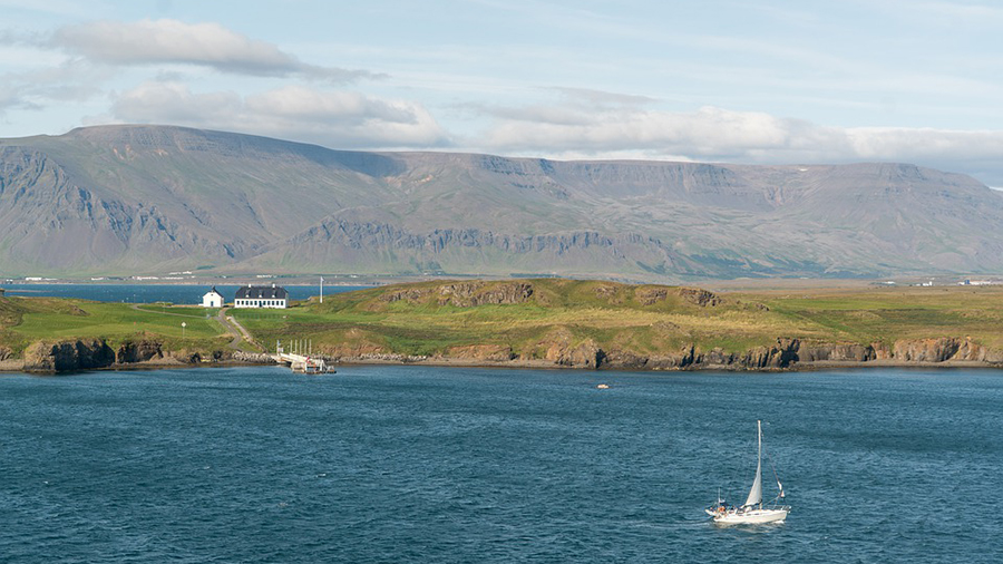 A sailboat is on a body of water near a small coastal area with cliffs and a few buildings, set against a backdrop of large, rugged mountains under a partly cloudy sky. The personnel on board seem well-organized, showcasing excellent people management skills.
