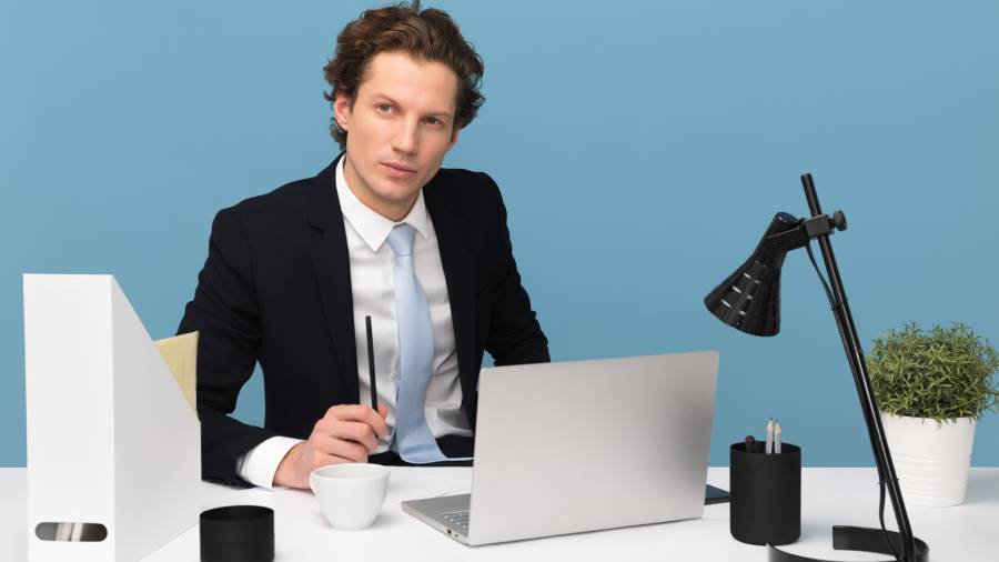 A man in a suit working at a desk with a laptop, notebook, and office supplies on a white surface against a blue background, focused on Human Resources tasks.