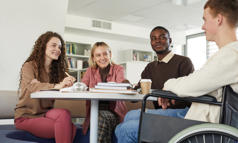 Four people are sitting around a table, engaged in conversation, with books and papers spread out in front of them. One person is in a wheelchair. They appear to be discussing recruiting strategies for individuals with a criminal conviction in a study or casual meeting setting.