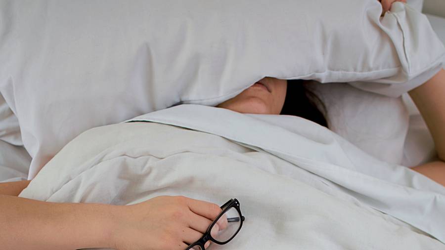 A person lies in bed with a pillow covering their face, holding a pair of glasses in one hand, perhaps contemplating the latest HR personnel reports.