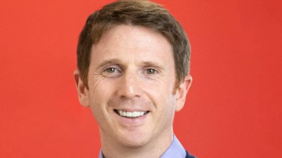 A man with short brown hair is smiling against a red background. He is wearing a light blue shirt and a dark sweater, embodying the approachable demeanor often valued in Human Resources.