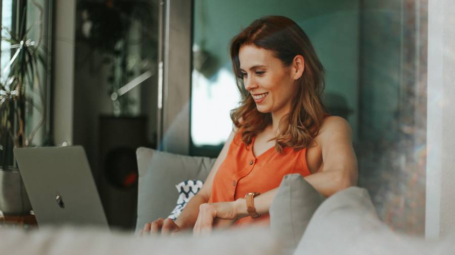 A woman sits on a sofa using a laptop, smiling and looking at the screen. Dressed in an orange sleeveless top, she appears engrossed in something related to People Management.