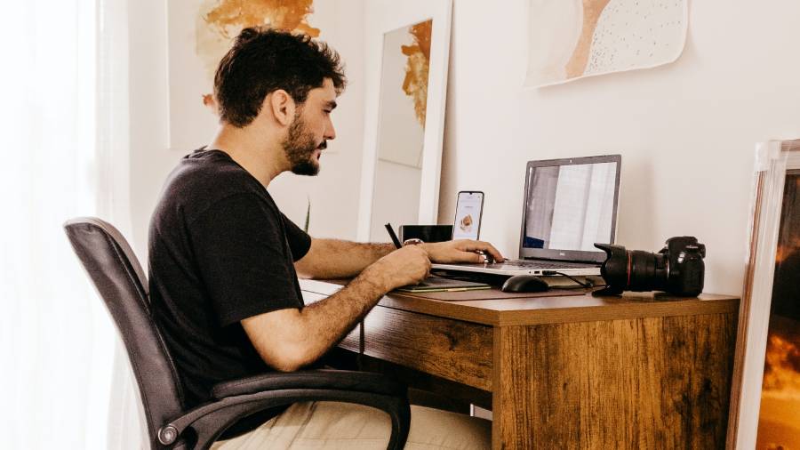 A man sits at a wooden desk using a laptop, with a smartphone and camera also on the desk. In his home office setting, he navigates through HR software, effectively managing personnel records alongside his other tasks.