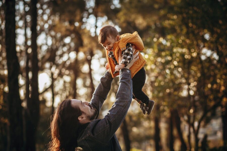 A man holds a toddler high in the air, with a wood in the background.