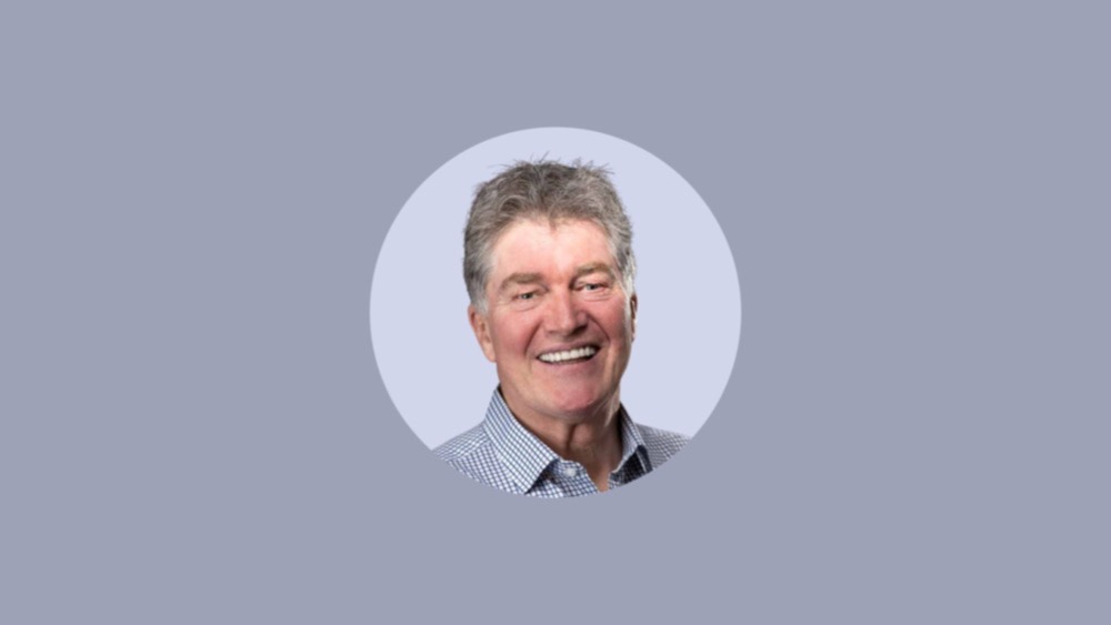 A smiling man with gray hair wearing a checkered shirt, embodying the return-to-office spirit, is centered in a circular frame against a light gray background.