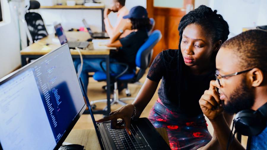 Two black employees at a desk are focused on a computer screen. One points at the screen while the other looks thoughtfully. Others work at desks in the background.