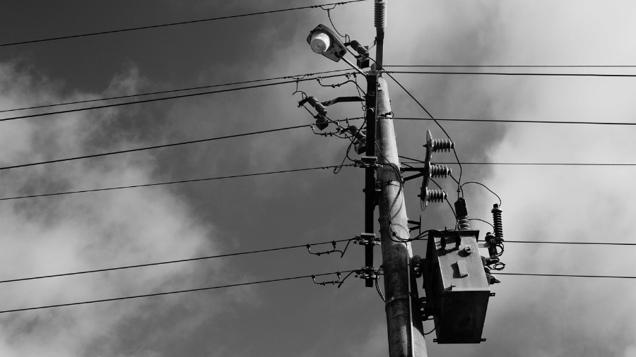 A black and white image of a utility pole with power lines and a street light against a cloudy sky, evoking the somber mood of the big switch off.