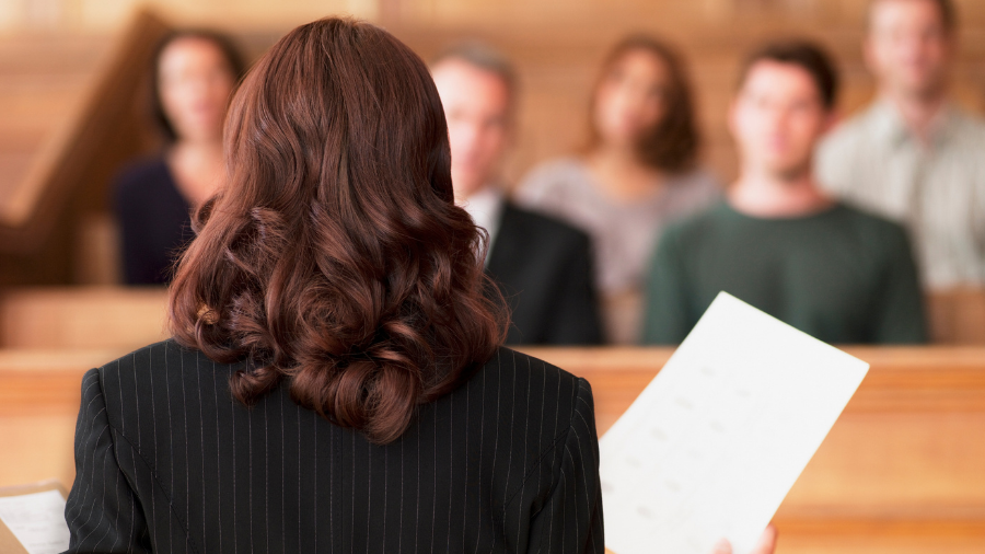 A person in a striped suit, seen from behind, holds documents while addressing four individuals seated in a courtroom setting, exemplifying the intersection of the legal profession and mental health advocacy.