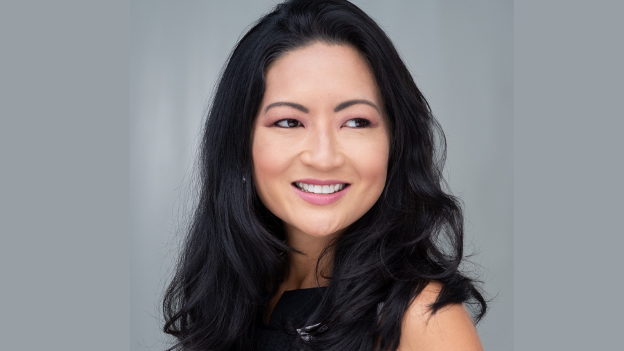 A woman with long black hair smiles and looks to the side. She wears a sleeveless black top and is set against a plain gray background, embodying diversity with her unique style and presence.