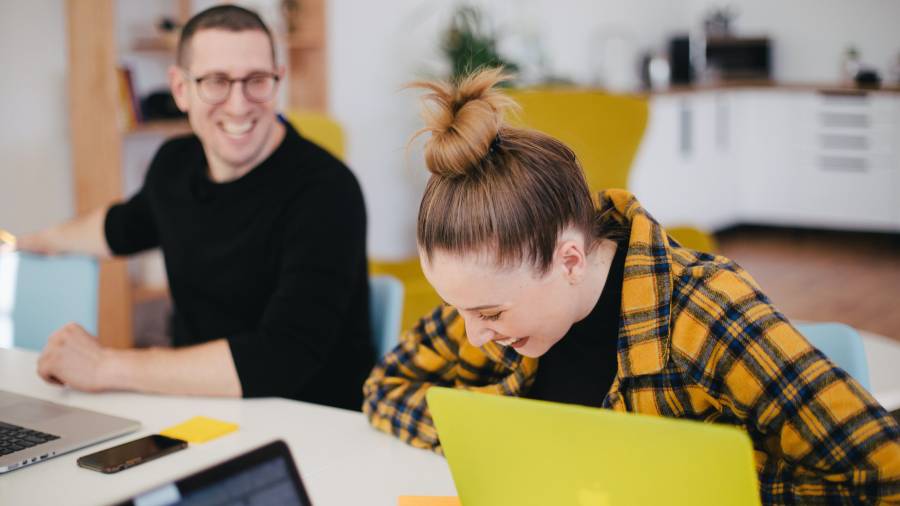 Two people sitting at a table, laughing and working on laptops. The person on the right has a yellow laptop and is wearing a yellow plaid shirt. The background shows a kitchen area, making for a relaxed atmosphere perfect for discussing people management strategies in human resources.
