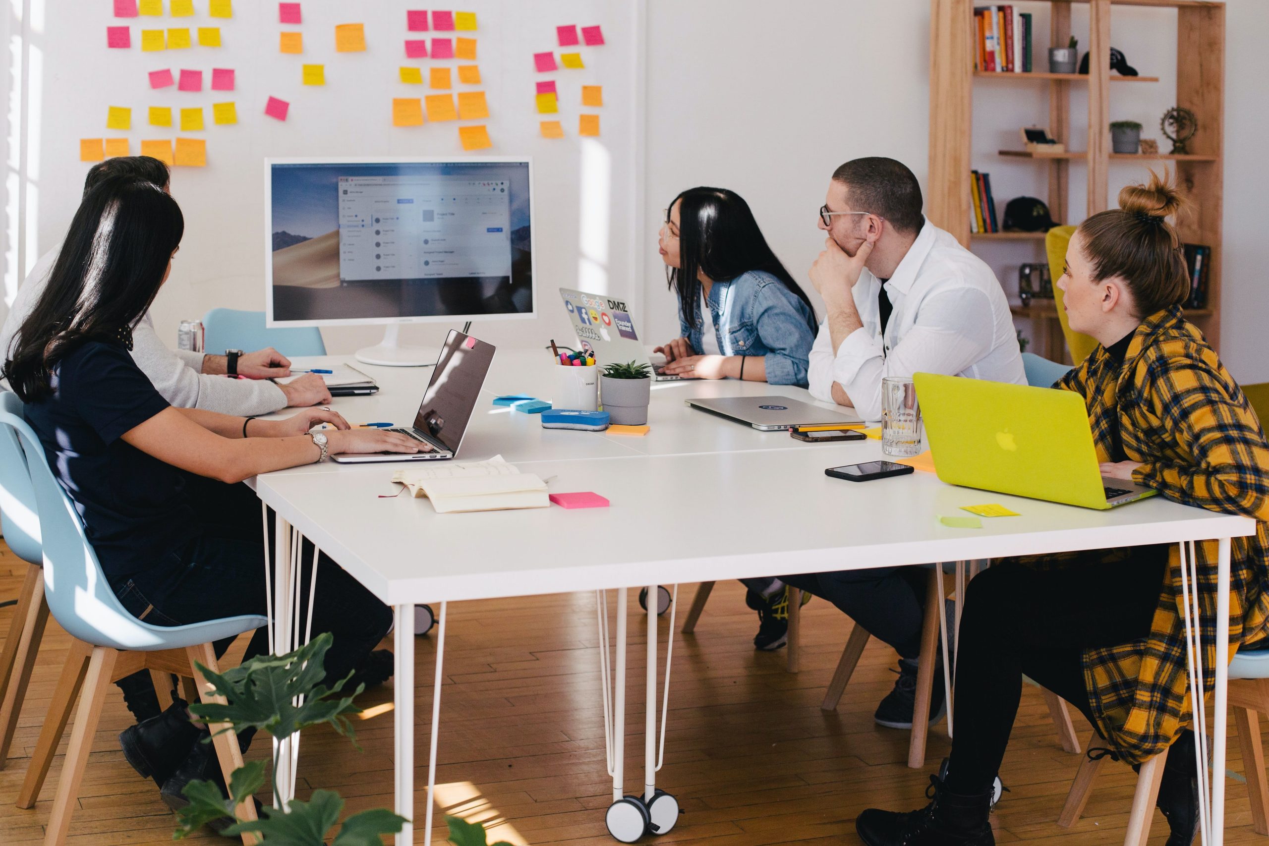 A group of five people sitting around a table in a meeting room, each with a laptop, is facing a large monitor displaying a presentation on candidate supply. Colorful sticky notes are on the wall in the background.
