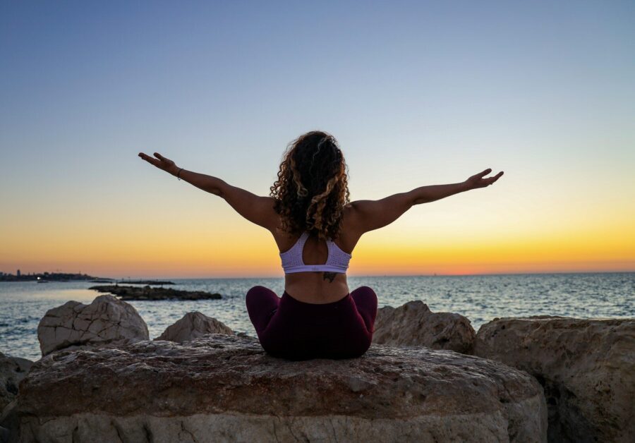 A woman sitting in front of the ocean at sunrise with her arms outstreched.
