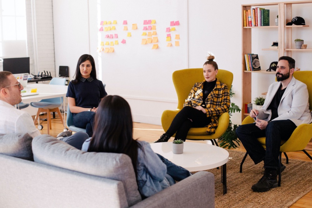 A group of five people sits in an office setting, engaged in a discussion about wellbeing support. Sticky notes are on the wall in the background, and bookshelves are to the right. The line manager leads the conversation, ensuring everyone’s concerns are addressed.