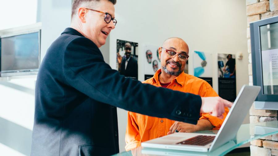 Two men discussing something on a laptop in a well-lit office. One man in a black jacket is pointing at the screen while the other in an orange shirt is smiling, engaging in a lively conversation about people management.