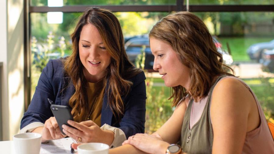 Two women sitting at a table, one showcasing something on her phone to the other. In the bright room with large windows in the background, their discussion appears focused on personnel matters and people management strategies.