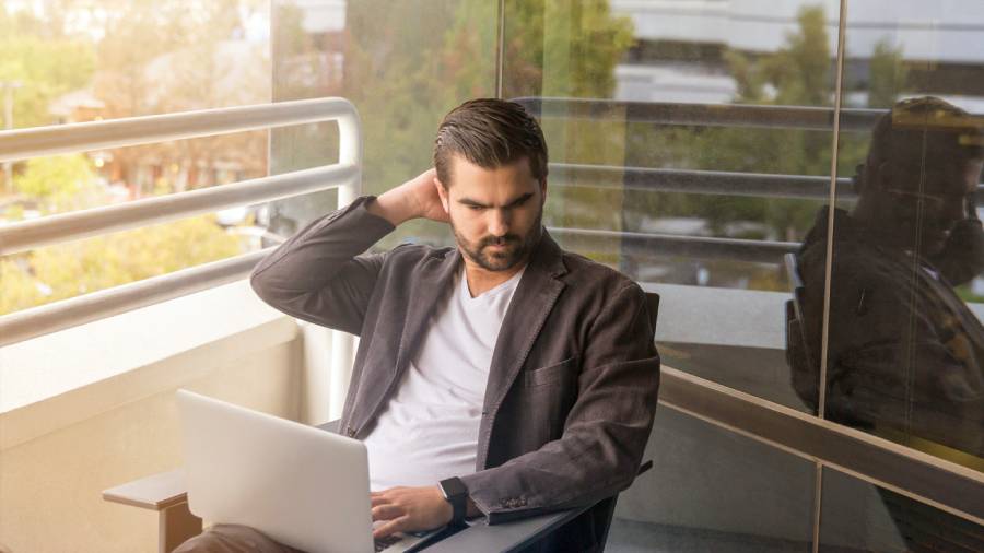 A man wearing a blazer and white shirt is sitting on a balcony, working on a laptop with HR documents, scratching the back of his head.