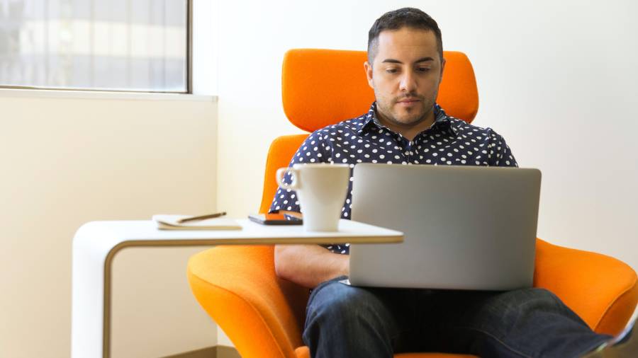 A man sits in an orange chair working on a laptop, possibly handling a Human Resources project. A white table beside him holds a smartphone, a notepad, and a coffee cup.