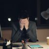A man in a suit sits at a desk with his head in his hands.