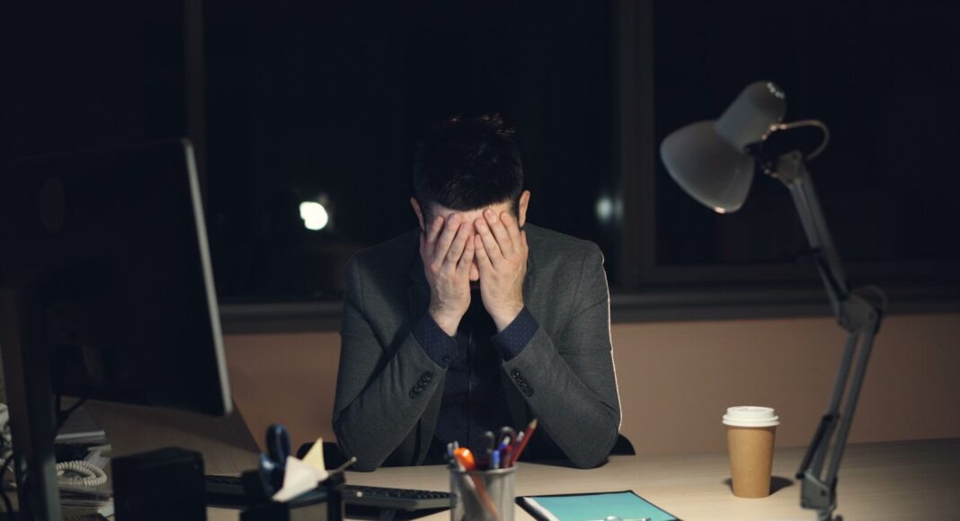 Photo by Vitaly Gariev A man in a suit sits at a desk with his head in his hands.