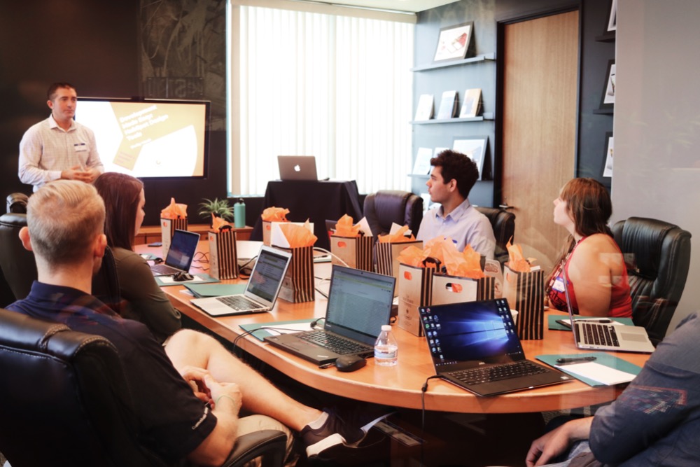 A group of people are seated around a conference table with laptops, listening to a man giving a presentation on disabilities in the workplace. Gift bags and water bottles are on the table.