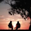 Silhouette of two people sitting on chairs near a tree at sunset.
