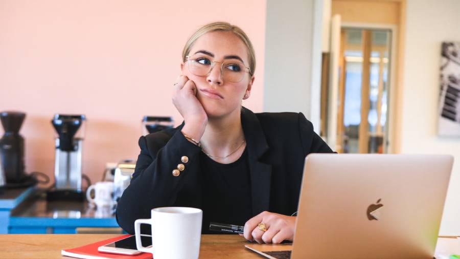 A woman wearing glasses sits at a desk with a laptop, coffee mug, and notebooks, resting her chin on her hand, appearing contemplative about Personnel management.