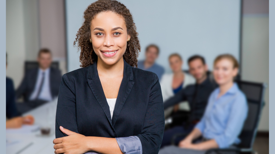 A person in a suit stands confidently with arms crossed in the foreground, exuding the essence of manager training, while a group of people sit at a conference table in the background, all facing the camera.