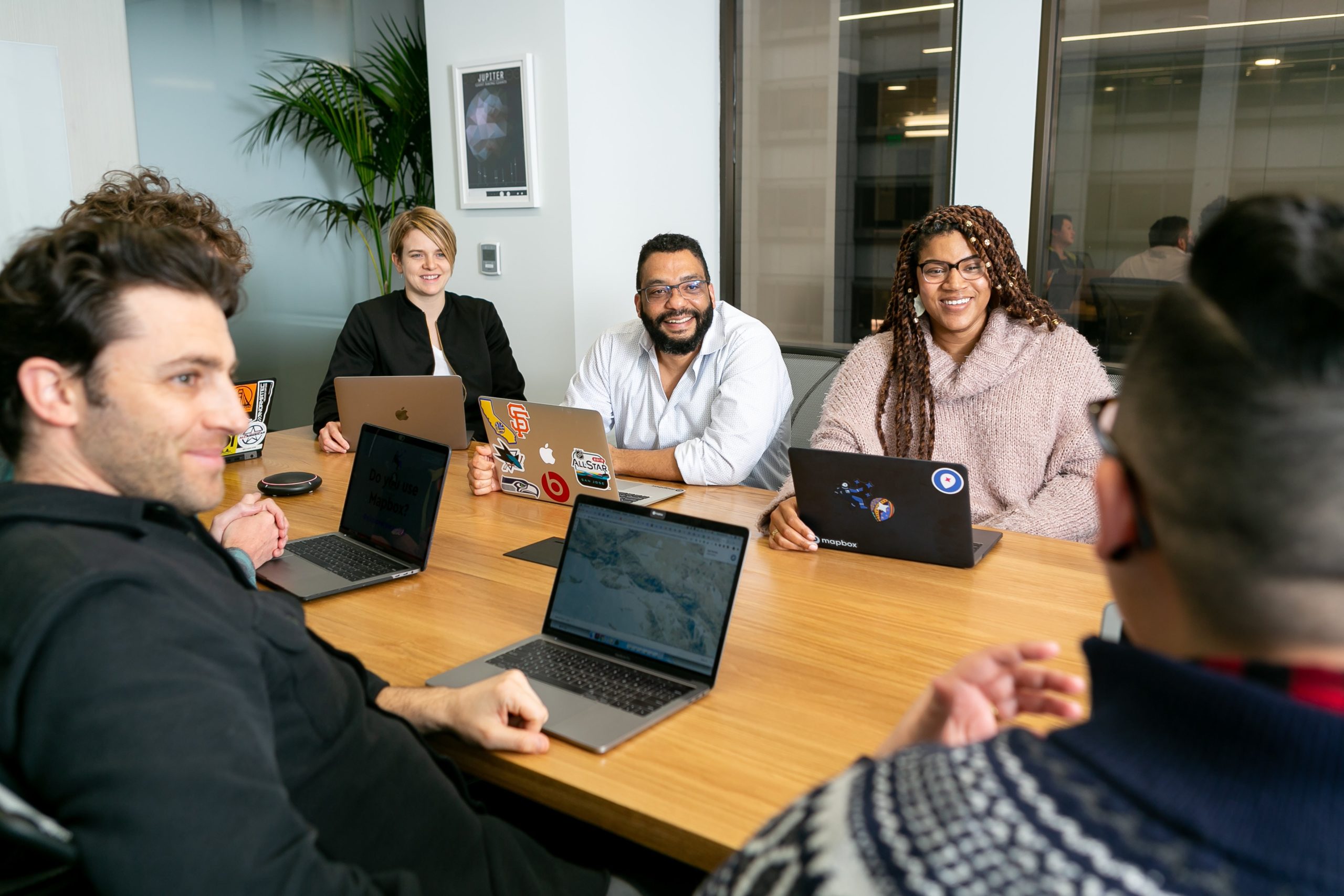 A diverse group of people, embodying the essence of hybrid working, sits around a table with laptops in a modern office setting, deeply engaged in discussion.