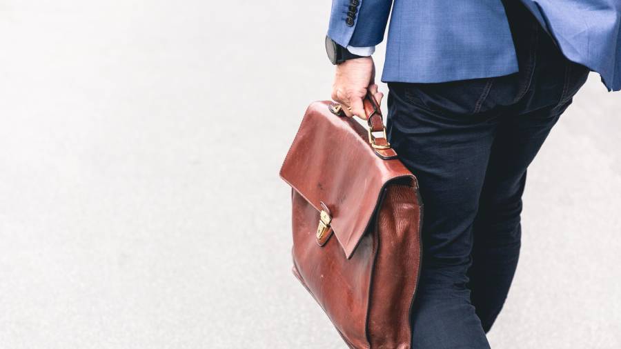 A person dressed in a blue blazer and dark pants is walking with a brown leather briefcase in their right hand, likely filled with important papers on people management and HR strategies.