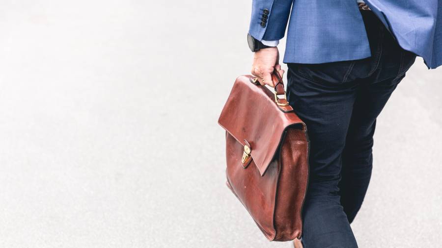 Person in a blue jacket walking on a concrete surface carrying a brown leather briefcase in their right hand, exemplifying professionalism in people management.