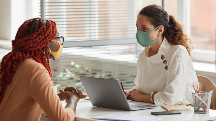 Two women wearing face masks sit at a table with a laptop, engaged in a conversation about hybrid working in a well-lit office setting.