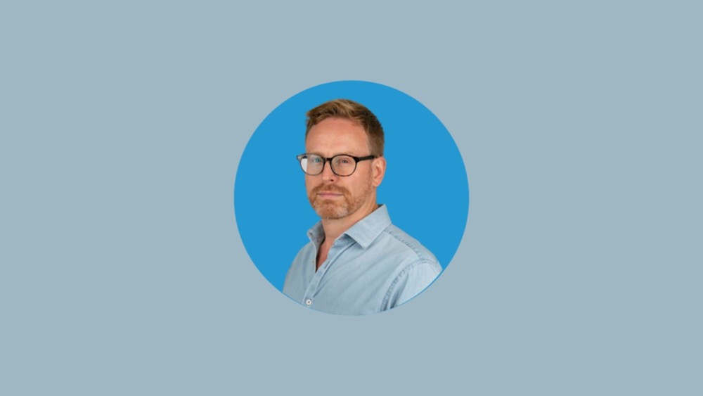A man with short hair and glasses, wearing a light blue button-down shirt, stands pensively against a blue background, perhaps reflecting on stress at work.