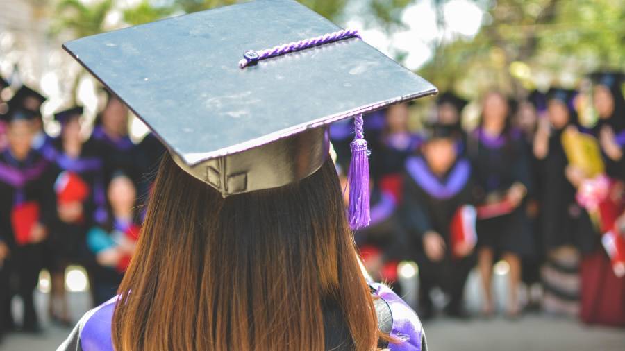 A graduate with long brown hair in a cap and gown stands facing a group of seated personnel in the background during a graduation ceremony.