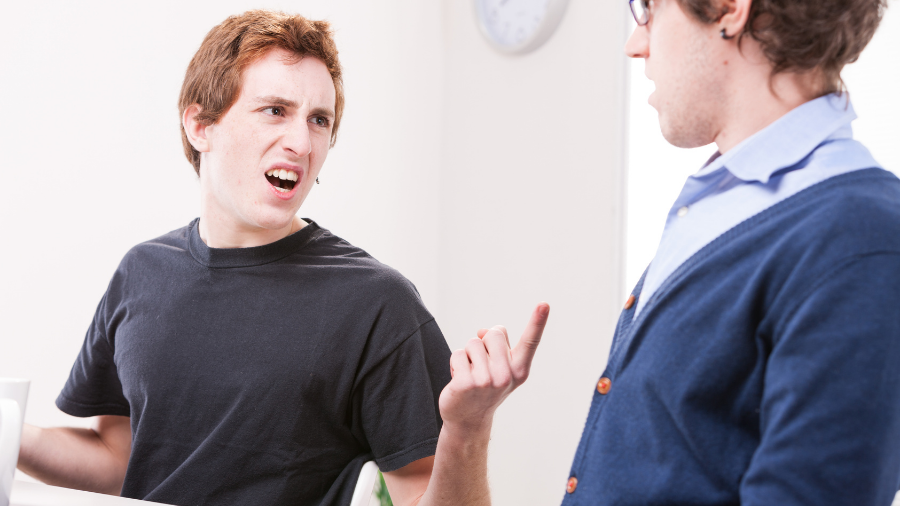 Two young men are in a heated discussion indoors. One on the left with a black t-shirt is gesturing and appears upset, evidently speaking about workplace sexism, while the other on the right in a blue sweater is responding. Two young men are in a heated discussion indoors. One on the left with a black t-shirt is gesturing and appears upset, evidently speaking about workplace sexism, while the other on the right in a blue sweater is responding.
