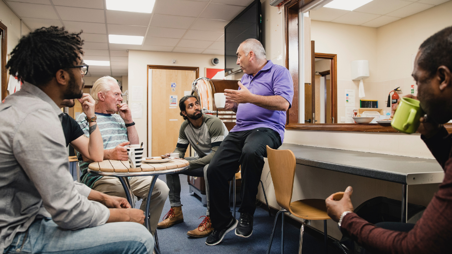 Five men are engaged in a conversation in a casual indoor setting with coffee cups. The man in the purple shirt appears to be leading the discussion, possibly addressing topics like workplace dynamics or menopause, while others listen attentively.