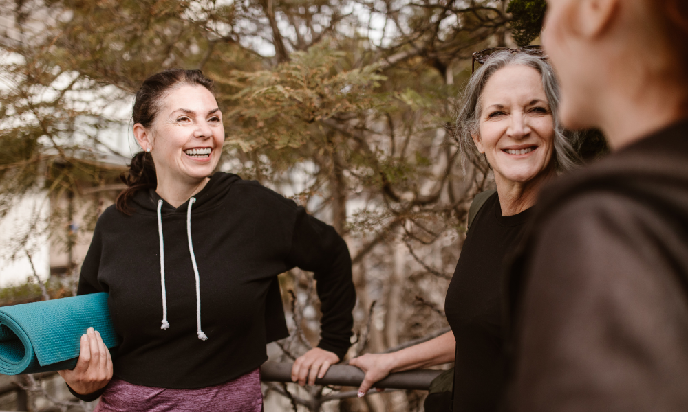 Three women in outdoor attire are smiling and conversing, with one holding a yoga mat. Trees are visible in the background, creating a serene setting perfect for discussing menopause support.