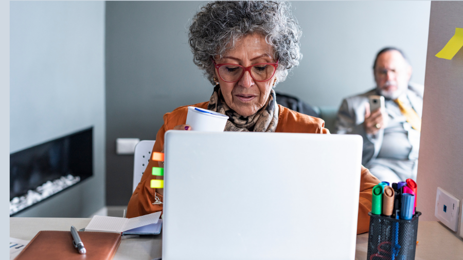 An elderly woman with curly gray hair and red glasses sits at a desk with a laptop, holding a disposable coffee cup. A man in a suit sits in the background looking at his phone. Office supplies are scattered on the desk, painting an everyday scene of menopausal life at work.