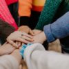 A group of individuals with colorful sweaters place their hands together in a circle, showing unity, teamwork, and promoting mental health.
