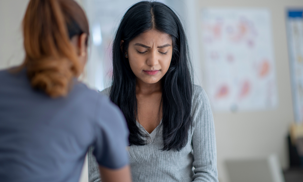 A woman in a gray sweater sits with a pained expression while a person in scrubs attends to her in a clinical setting, highlighting the importance of employee mental health.