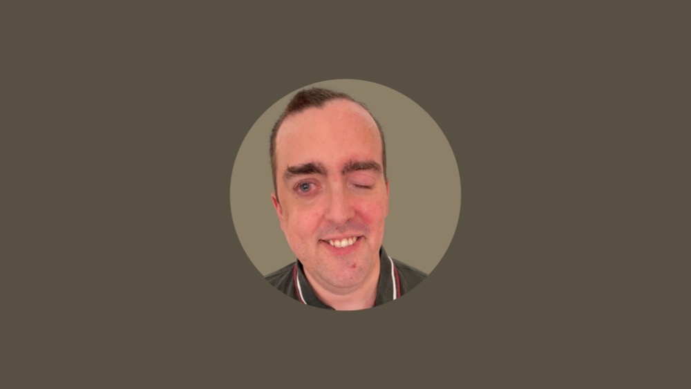 A man with short dark hair and a smile stands confidently. He is wearing a dark collared shirt, embodying both professionalism and approachability. The background is a muted grey color, subtly emphasizing the importance of accessibility practices in design choices.